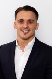 Professional headshot of a smiling man with dark hair wearing a navy blazer and white dress shirt against a light gray background